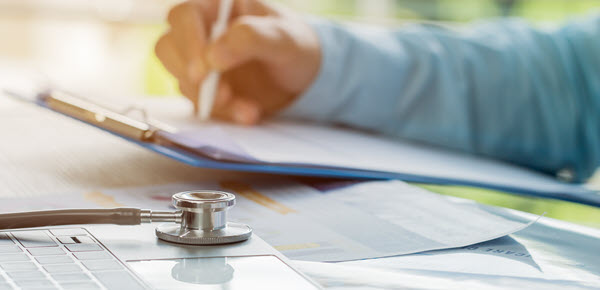 Doctor writing, with a laptop and stethoscope in the foreground