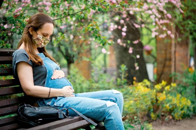 Pregnant vaccination A pregnant woman wearing a cloth facemask sits on a park bench holding her belly.