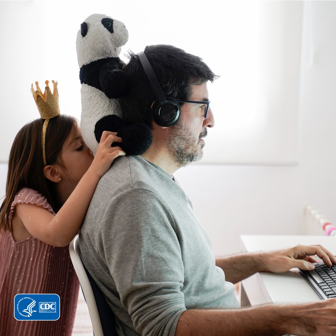 Daughter standing behind her father holding a stuffed panda on his shoulders as he is on the computer.