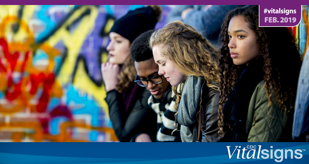 Three teenage girls and one teenage boy sit outside with graffiti in the background.