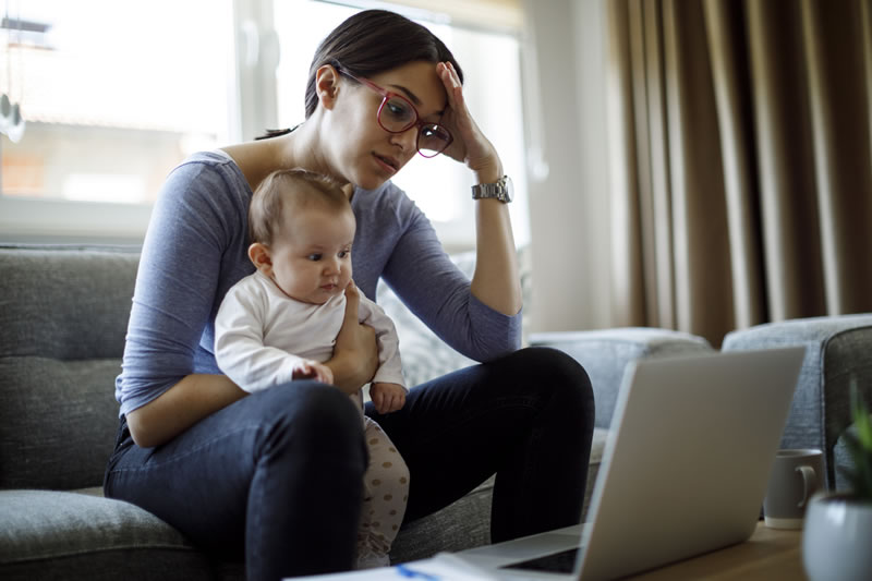iStock-1220413419 woman sitting on sofa holding baby, looking at laptop