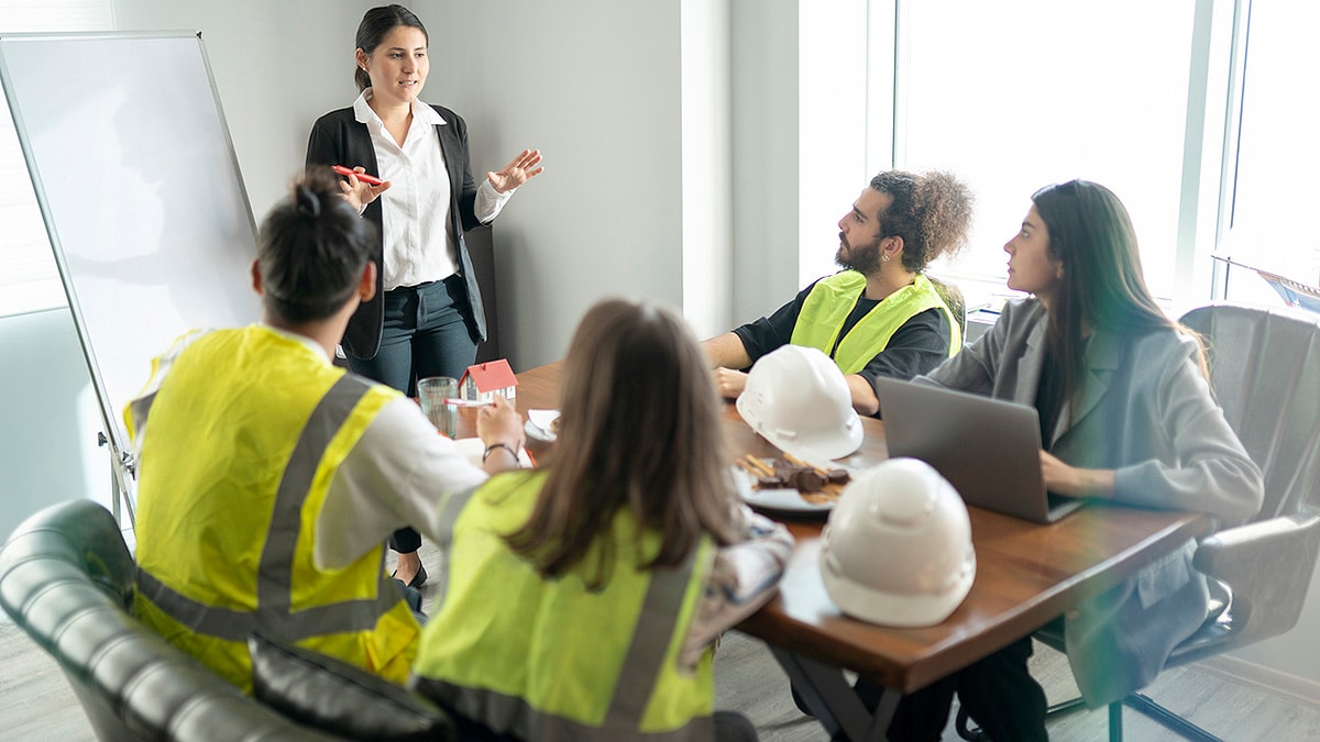 Workplace Health Train the Trainer Businesswoman standing at dry erase board training three workers in hardhat gear and one woman in business attire.